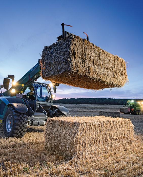 Verreiker stapelt grote balen stro op het veld bij zonsondergang